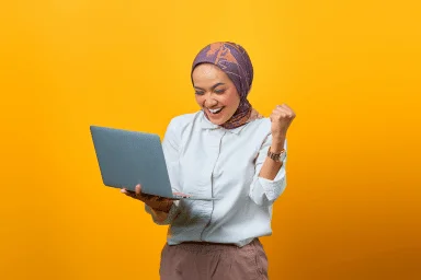 Woman with scarf in her hair holding a laptop while celebrating with her sawn fist.